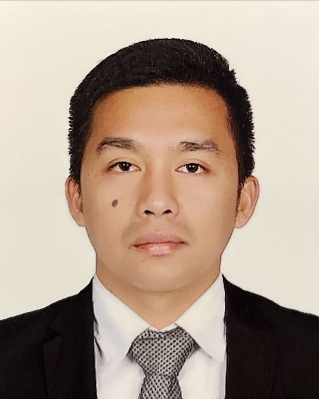 Karl Jose Buena, head and shoulders, in a dark suit, white shirt and grey patterned tie, against a plain white background.